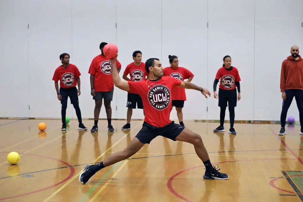 UCW MBA Games dodgeball team member in red shirt and black shorts prepares to throw the ball as six team members look on.