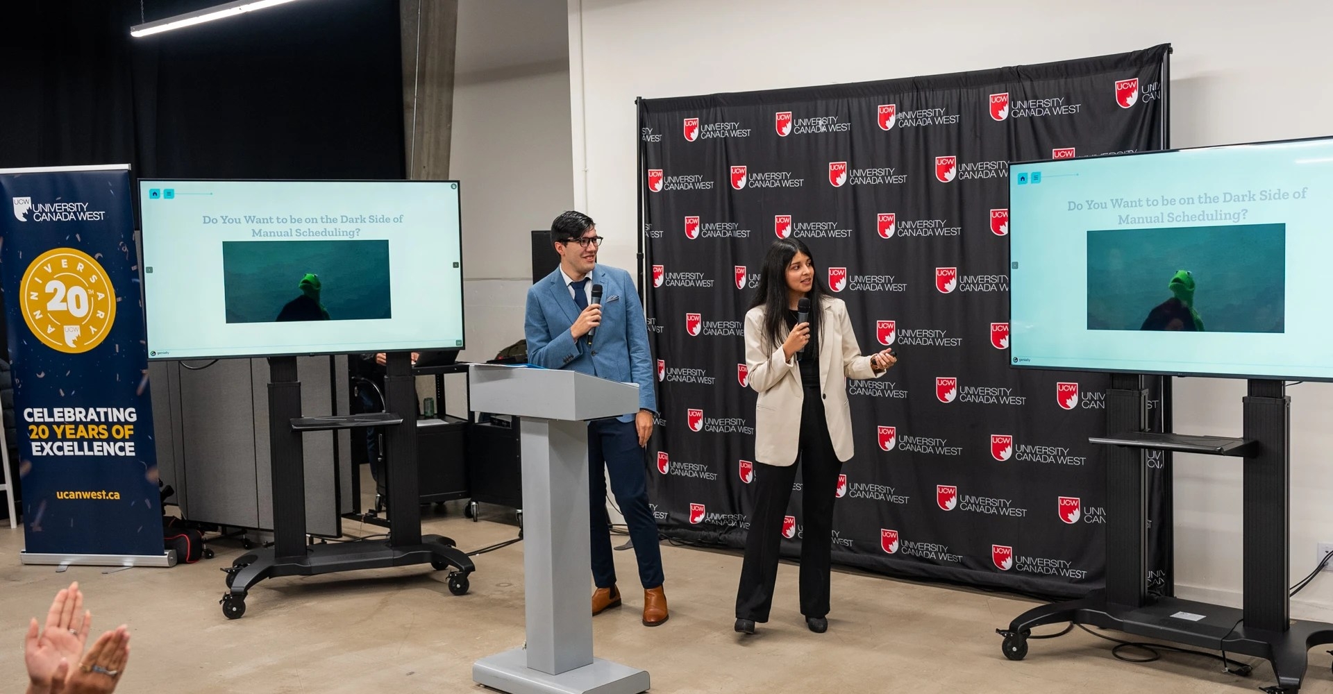 A male and female student are holding microphones talking to a slide that says "Do you want to be on the dark side of manual scheduling" as they pitch their business idea.